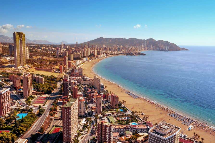 A view of Levante Beach, with golden sand stretching along it, next to a city of skyscrapers under a clear blue sky.