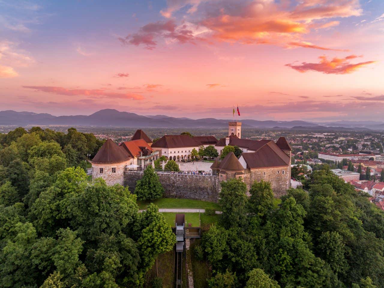 Il castello di Lubiana in cima a una verde collina al tramonto, il cielo presenta sfumature arancioni, rosse e rosate, mentre all'orizzonte di vede il profilo delle montagne