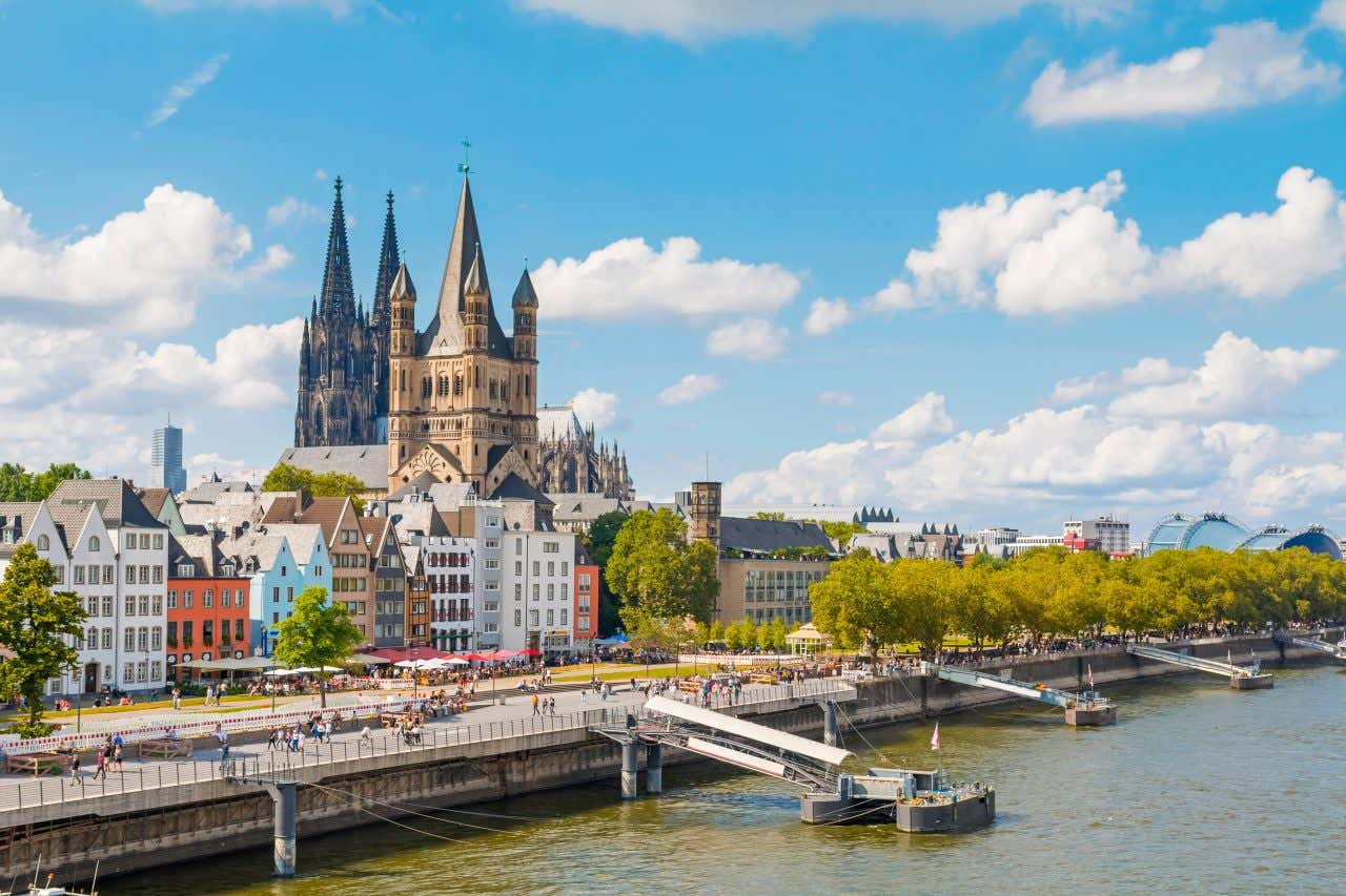 The Hohenzollern Bridge along the waterfron, lined with traditional, colorful houses, the Cologne Cathedral visible in the background on a sunny day