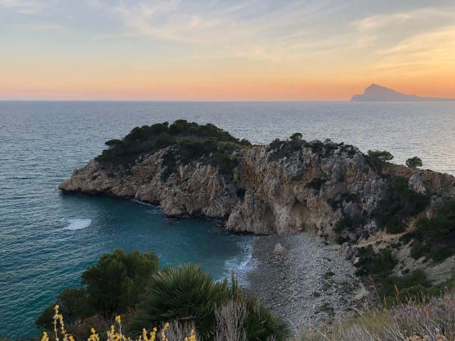 An aerial view of the El Mascarat Beach, with a rocky outlet, seen at sunset.