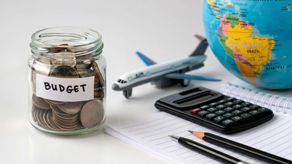 Un quaderno con penne e matite, un barattolo di vetro pieno di monete, una calcolatrice, il modellino di un aereo e un mappamondo poggiati su un tavolo a simboleggiare la pianificazione per un viaggio low cost