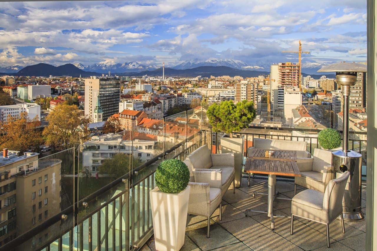 Una terrazza con un tavolino e alcune sedie di design. Sullo sfondo la città di Lubiana dall'alto e alcune montagne innevate all'orizzonte