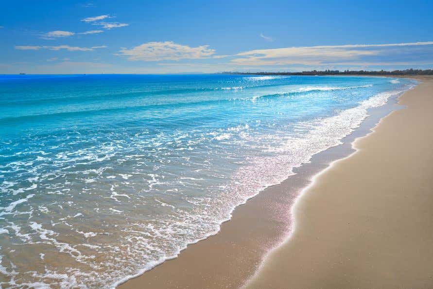 A view of Pinedo Beach, with waves crashing on the golden sand, under a blue sky with a few white clouds.