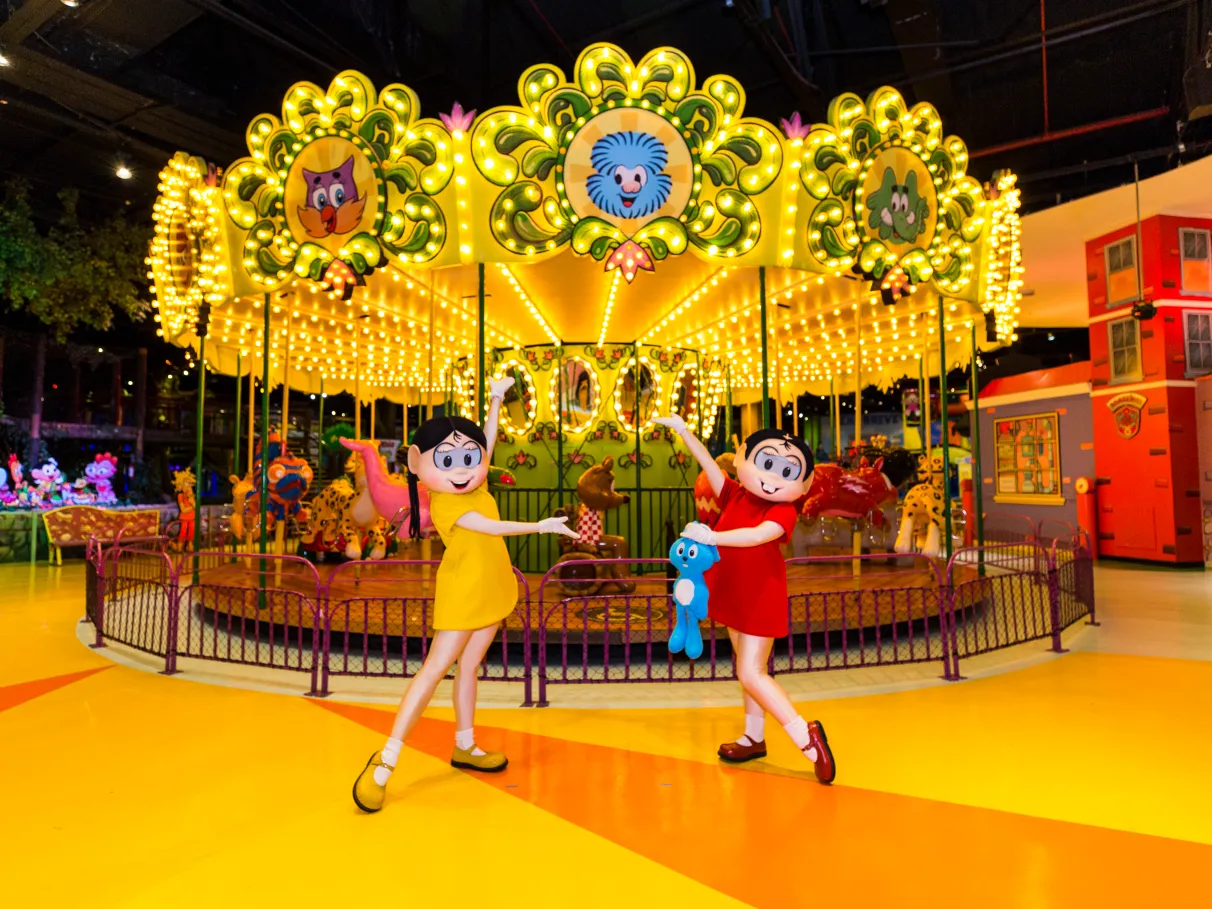 As personagens Magali e Mônica, segurando o Milu, da Turma da Monica posando para uma foto em frente a um carrossel iluminado
