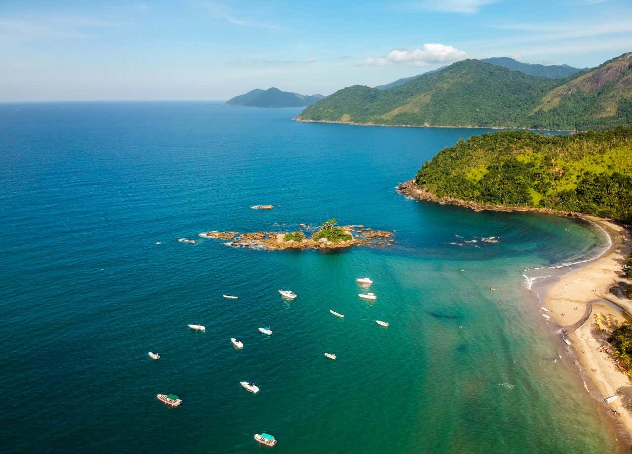Vista aérea de uma praia com águas turquesa com vários barcos e montanhas ao fundo