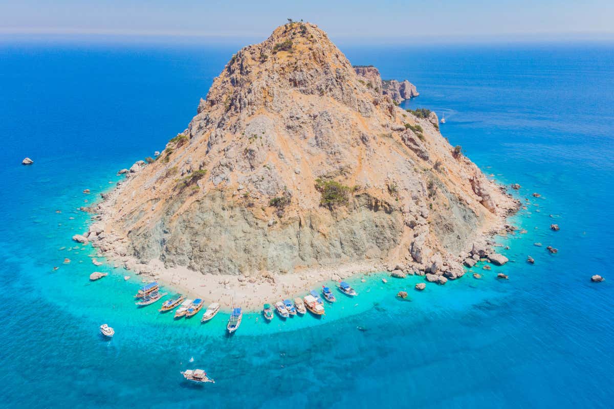 Vista aérea de una isla desértica rodeada de aguas turquesas y unos barcos fondeados en su costa