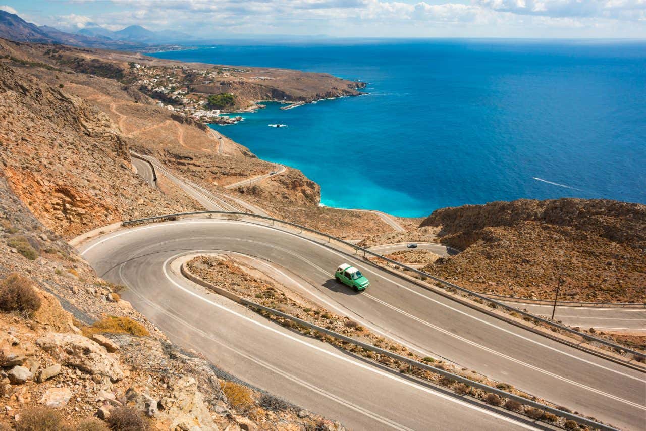 Vista aerea di una strada costiera di Creta e un'auto che sfreccia su un tornante, il mare cretese sullo sfondo