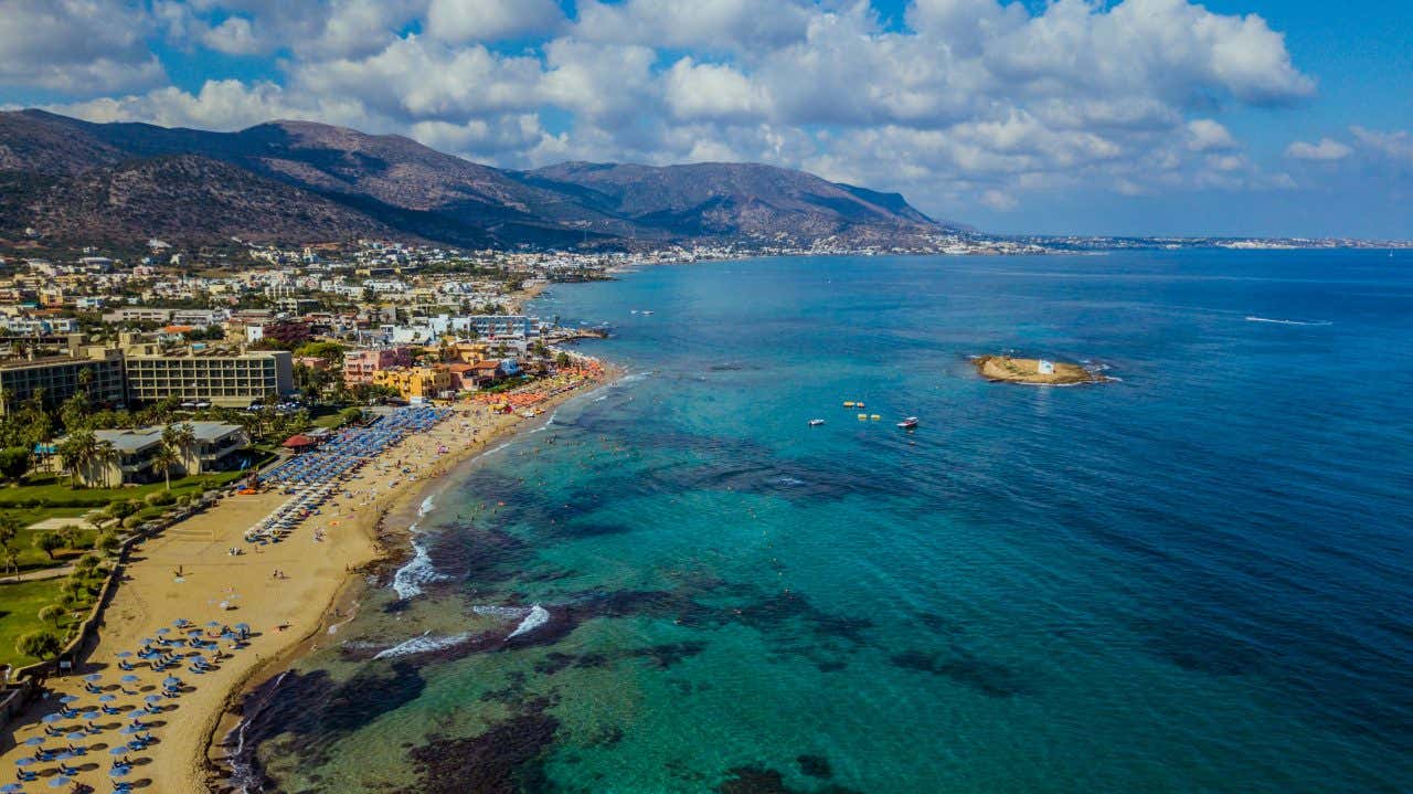 Vista aerea di una lunga spiaggia sabbiosa con ombrelloni e lettini, affacciata su un mare turchese e trasparente, con una cittadina costiera e montagne sullo sfondo, e una piccola isola con una struttura bianca al largo