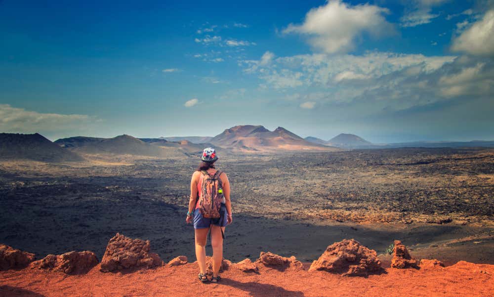 Un'escursionista in vestiti estivi fotografata di spalle mentre osserva un paesaggio roccioso con alcune vette spoglie da un'altura pianeggiante fatta di terra rossiccia