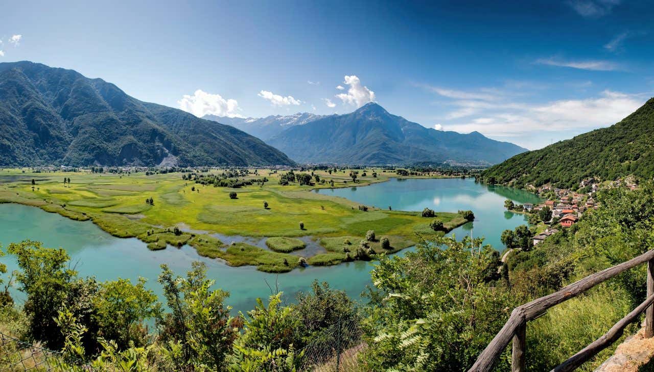 Panoramica del Pian di Spagna in Lombardia, con prati verdi, corsi d'acqua intrecciati e montagne sullo sfondo, fotografati da un punto panoramico rialzato con vegetazione in primo piano e un villaggio sulla destra