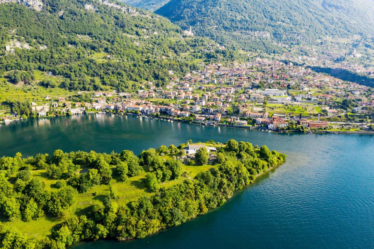 Veduta aerea dell'Isola Comacina sul Lago di Como in una giornata soleggiata, circondata da acque blu e da una fitta vegetazione, con il paese di Ossuccio e le montagne verdeggianti sullo sfondo