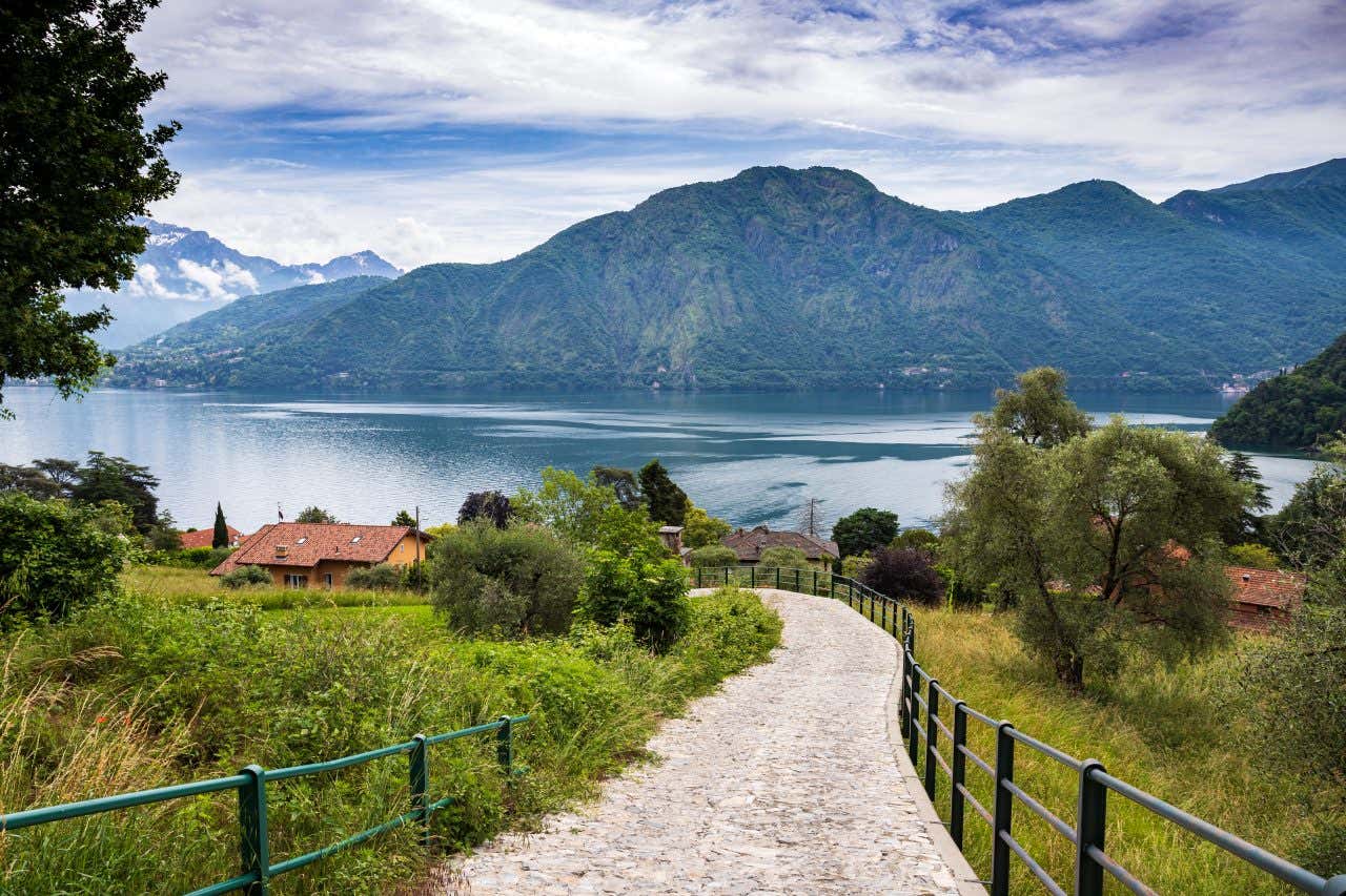 Sentiero acciottolato che scende verso il Lago di Como, circondato da vegetazione, ulivi e case rustiche, con le montagne verdeggianti sullo sfondo sotto un cielo parzialmente nuvoloso