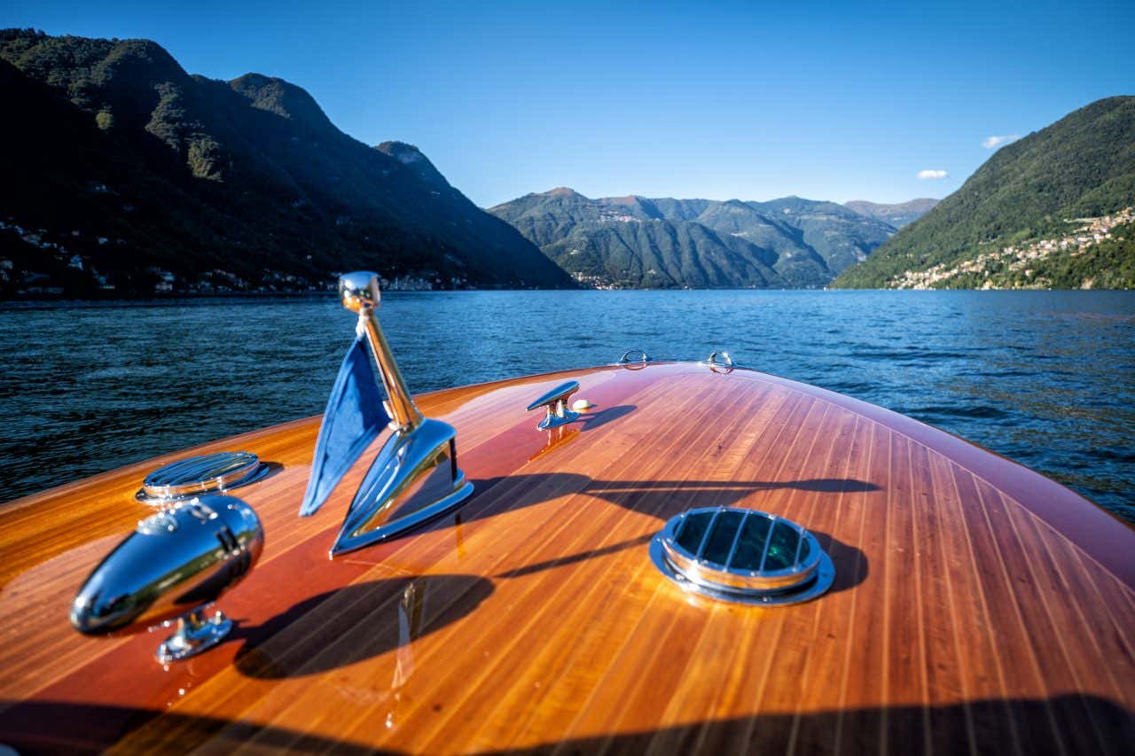 La prua di una barca di lusso in legno mentre naviga sul Lago di Como, all'orizzonte ci sono le vette di alcune montagne e piccoli villaggi su entrambe le rive del lago