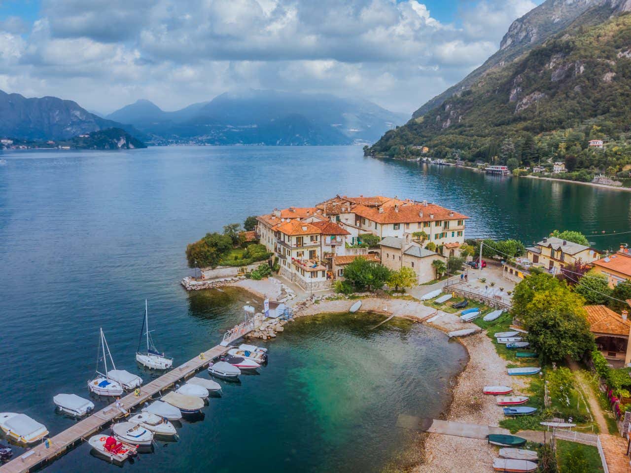 Veduta aerea di Lierna, pittoresco borgo sul Lago di Como, con case dai tetti rossi affacciate sull'acqua cristallina, piccola spiaggia e porto con barche ormeggiate, circondati da colline verdi e montagne sullo sfondo