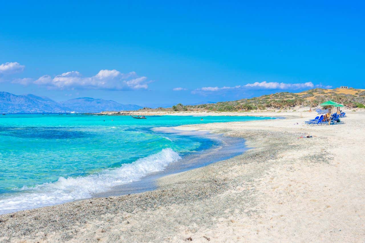 Una spiaggia di sabbia dorata bagnata da un mare di colore turchese intenso, sulla sabbia ci sono alcuni ombrelloni e si intravedono piccole porzioni di cespugli bassi