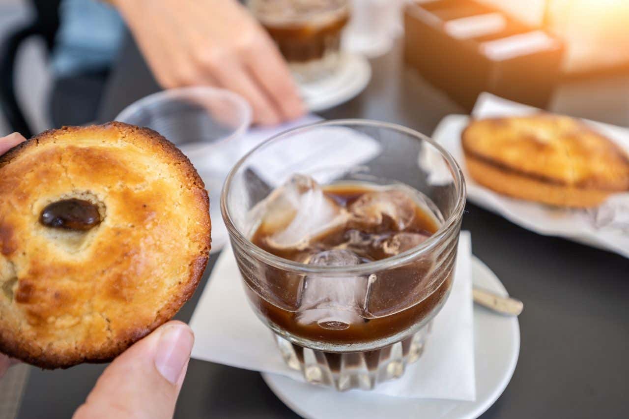 Un caffè con ghiaccio dentro un bicchiere di vetro al centro di un tavolino di un bar. In primo piano una mano tiene un pasticciotto ripieno, dolce tipico della tradizione leccese