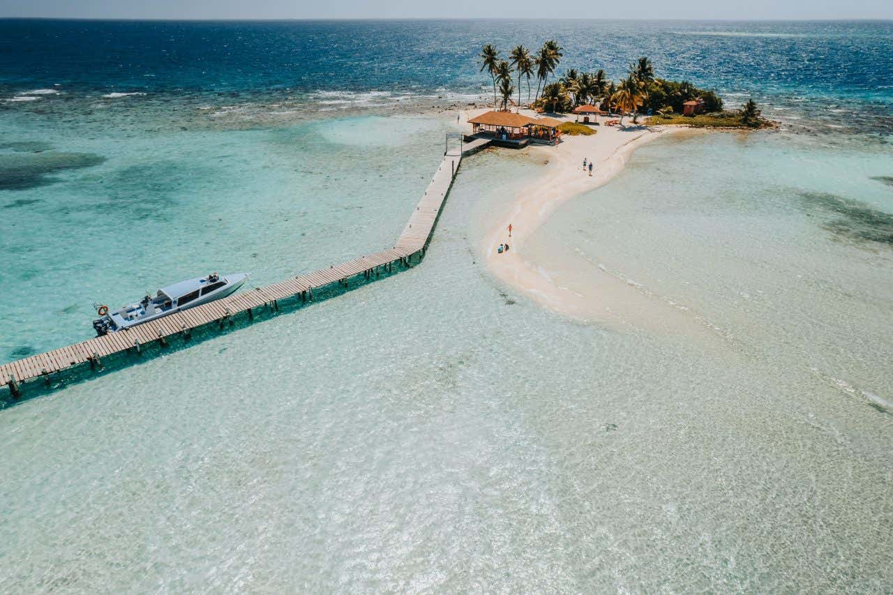 An aerial view of Goff's caye, with a boat docked at a wooden dock, and people walking along a sand bridge.