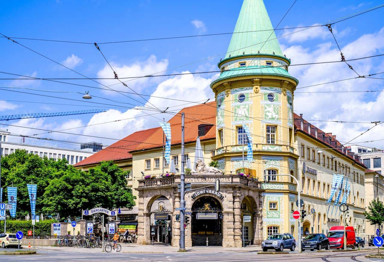 Fachada da Löwenbräukeller, um prédio amarelo com entrada de pedra em colunas e uma torre amarela com telhado verde