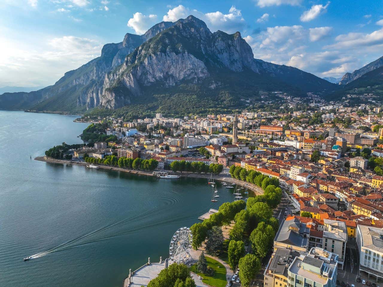 La città di Lecco affacciata sul lago di Como con un lungolago alberato, case di colore giallo e arancione e un alto e maestoso monte sullo sfondo in una giornata di sole