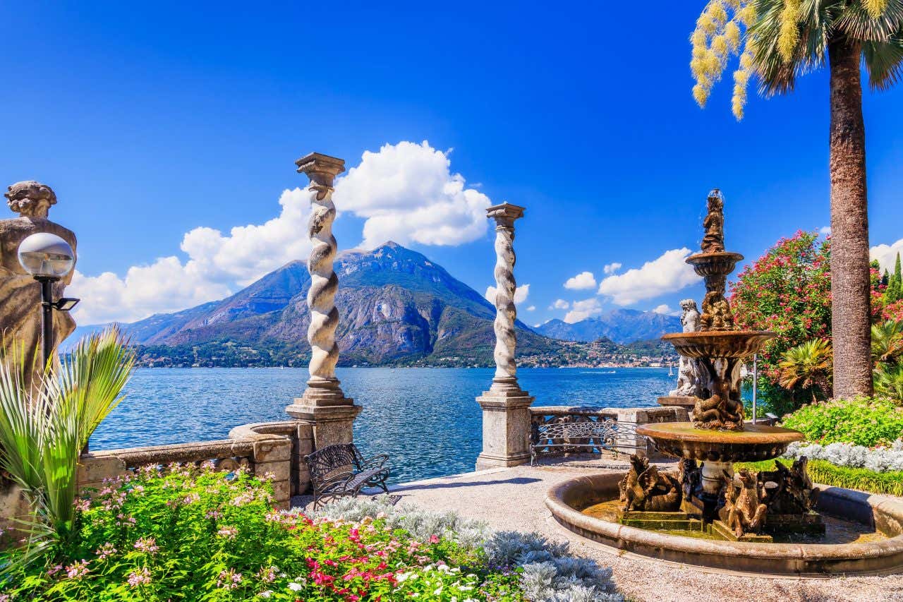Elegante giardino sul Lago di Como a Varenna, con colonne intrecciate in pietra, fontana decorativa, fiori colorati e una palma, vista panoramica sul lago e sulle montagne sotto un cielo azzurro e nuvole bianche