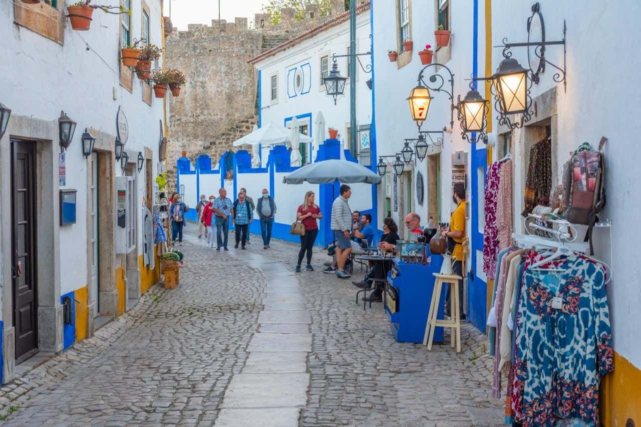 A cobblestone street in Óbidos, Portugal, lined with white buildings featuring blue and yellow painted details. People are walking, standing, and sitting at outdoor tables.