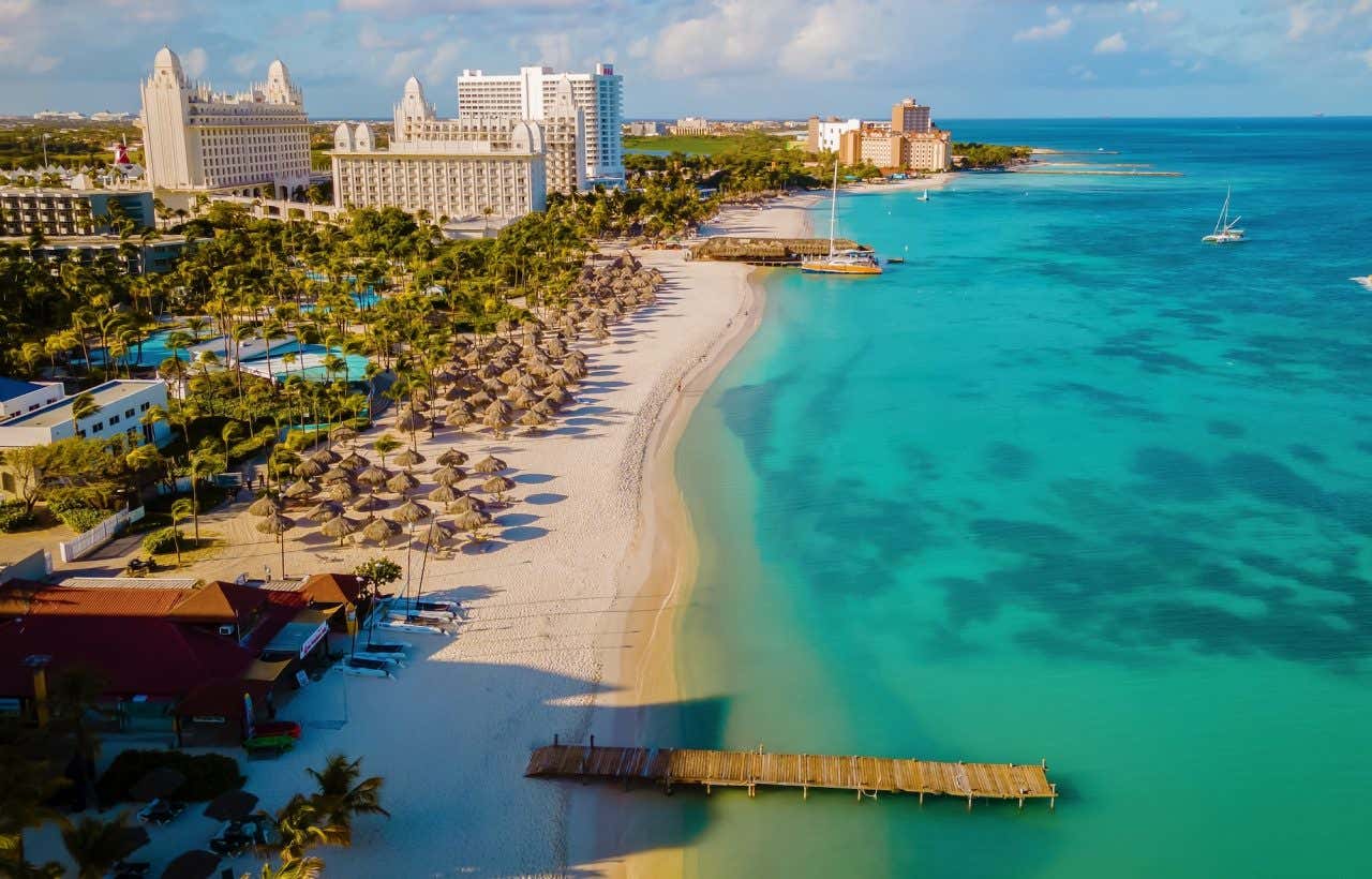 Vista aérea de uma praia com areia branca e águas turquesas com muitos guarda-sóis de palha e resorts ao fundo