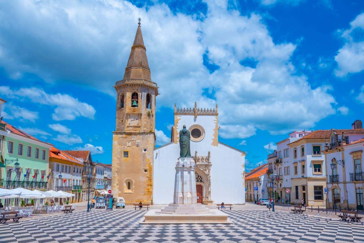 The Church of São João Baptista with a tall bell tower stands in a large, checkered town square in Tomar, Portugal. A statue is located in the center of the square, and other buildings are visible around the perimeter.