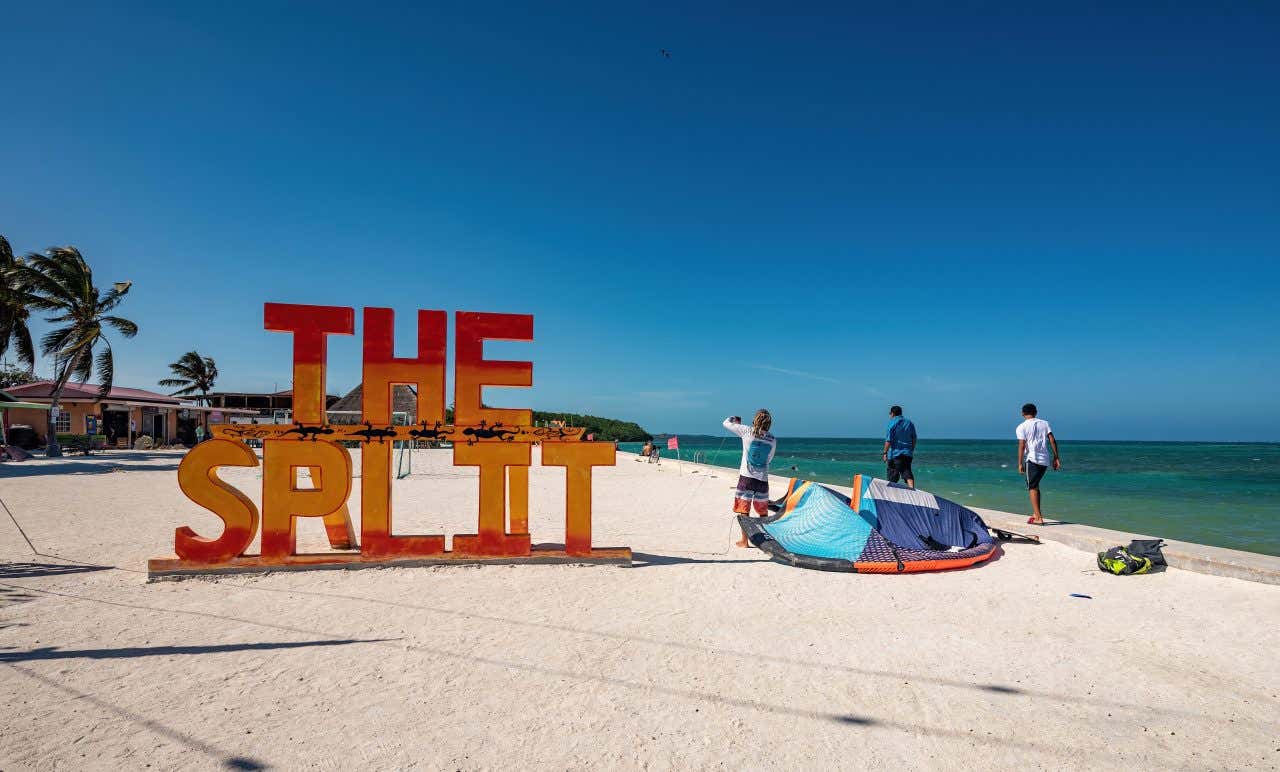 'The Split' sign written on The Split Beach in colorful red and yellow block lettering, with 3 people standing beside a tent to the right of the sign.