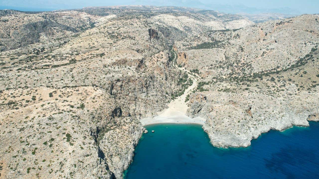 Veduta aerea di una spiaggia nascosta tra scogliere rocciose e paesaggio arido, con mare cristallino di colore blu intenso, situata in una baia appartata
