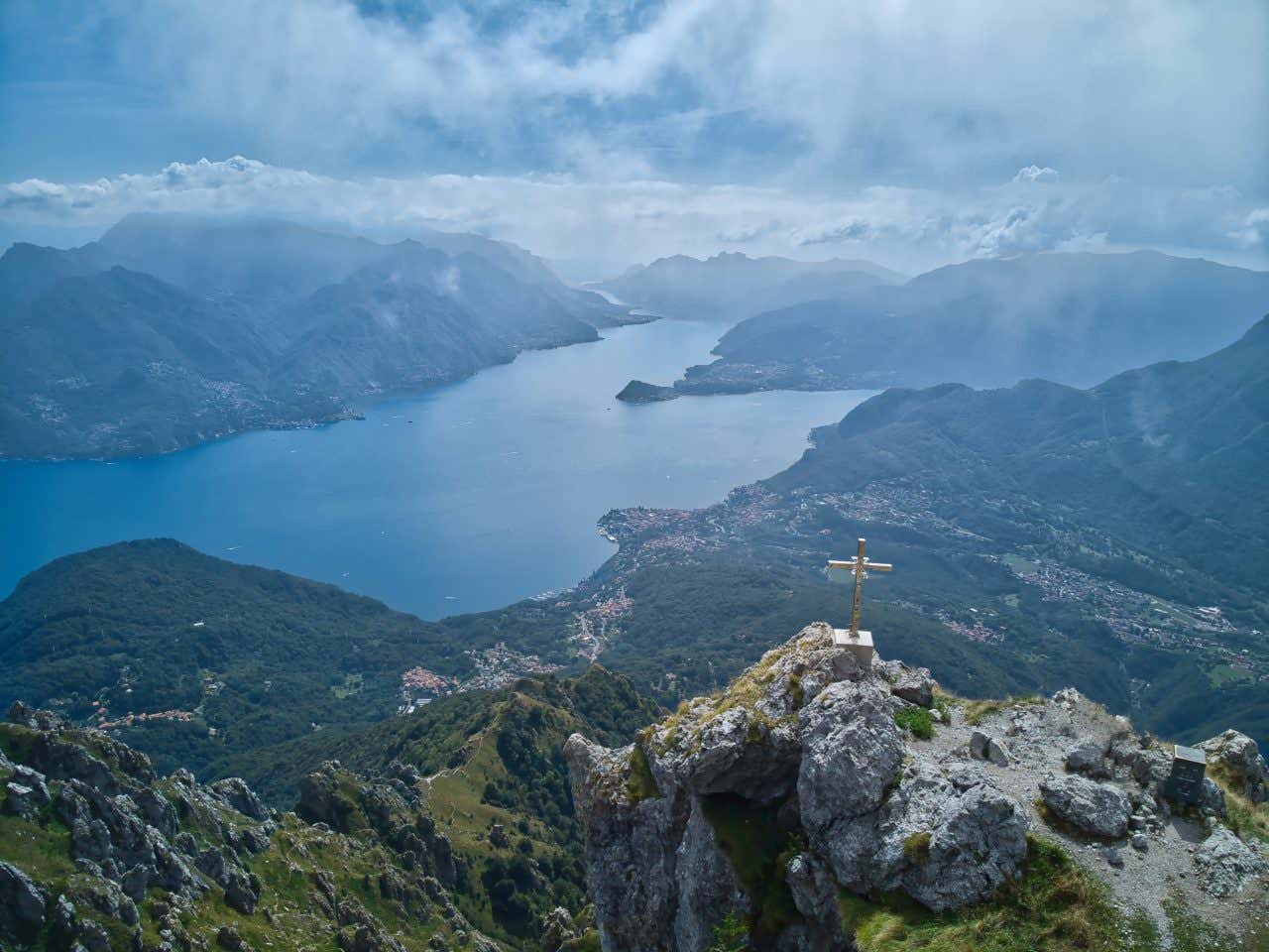 Vista del Lago di Como dall'alto di una cima montuosa segnalata da una croce di metallo. Si vedono i due bracci del lago che si separano all'altezza del promontorio di Bellagio