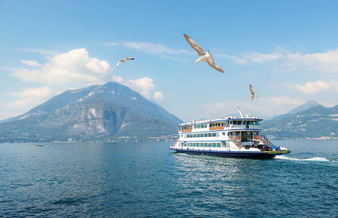 Battello in navigazione sul Lago di Como con partenza da Varenna, circondato da gabbiani in volo e montagne sullo sfondo in una giornata soleggiata