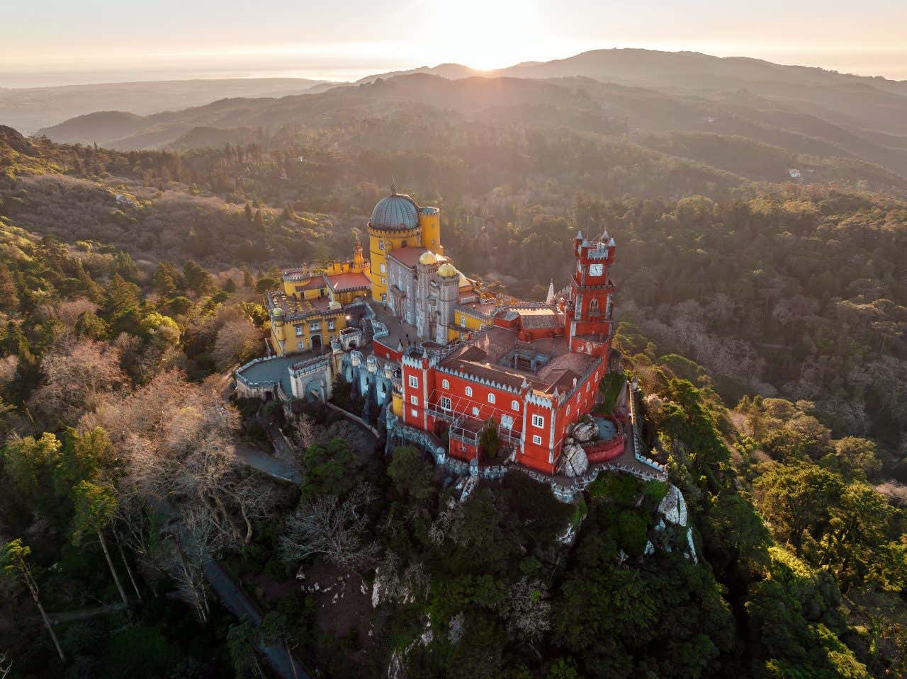 An aerial view of Pena Palace in Sintra, Portugal. The multi-colored castle is situated on a forested hilltop, with sunlight illuminating part of the palace and the surrounding landscape.