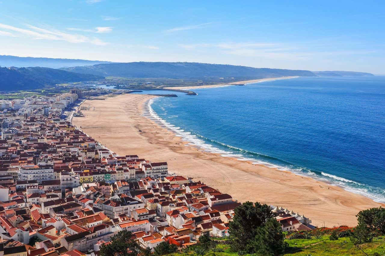 A high-angle view of Nazaré, Portugal, showing a long sandy beach curving along the ocean. A town with multiple buildings and terracotta rooftops is visible along the coast and extending up the hillside.