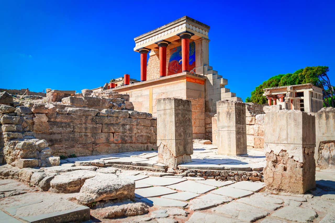 Vista dal basso del punto più famoso del palazzo di Cnosso a Creta in una giornata di sole. In primo piano ci sono i resti di mura, mentre i secondo piano, posti in cima a un muro di pietre chiare, c'è la ricostruzione di un colonnato rosso e affreschi colorati sulla parete retrostante