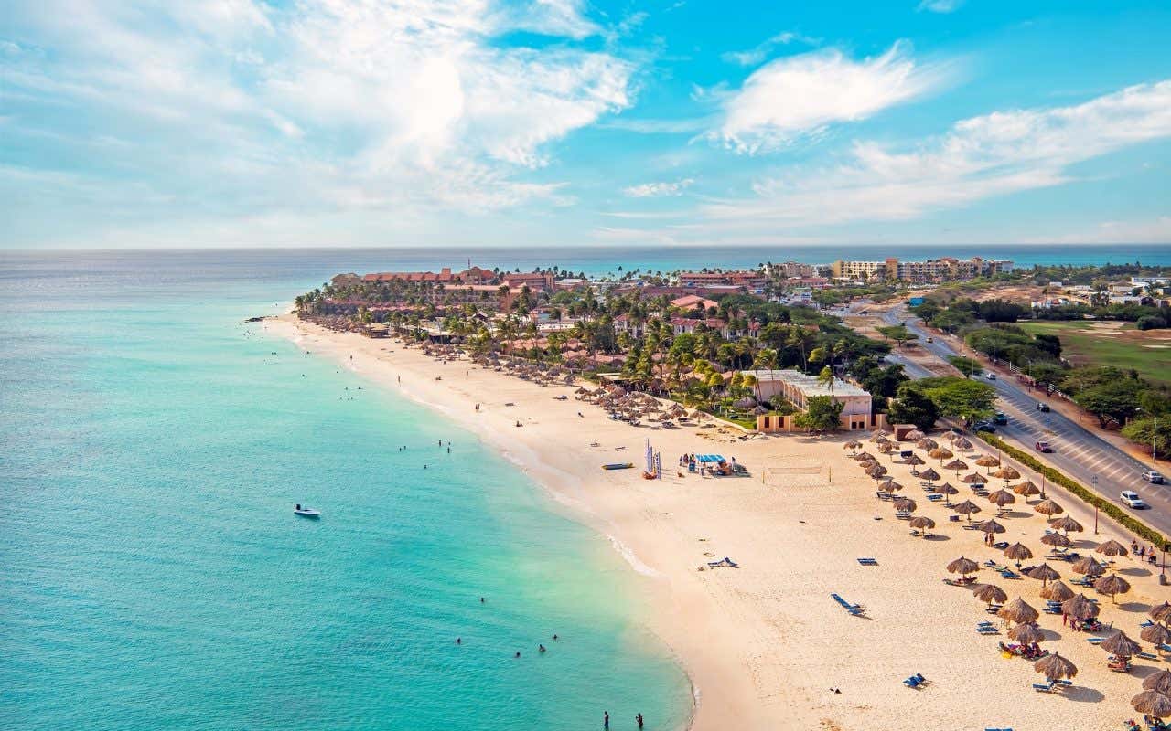 Vista aérea de uma praia com areia branca e águas turquesas com muitos guarda-sóis de palha e casas e árvores ao fundo