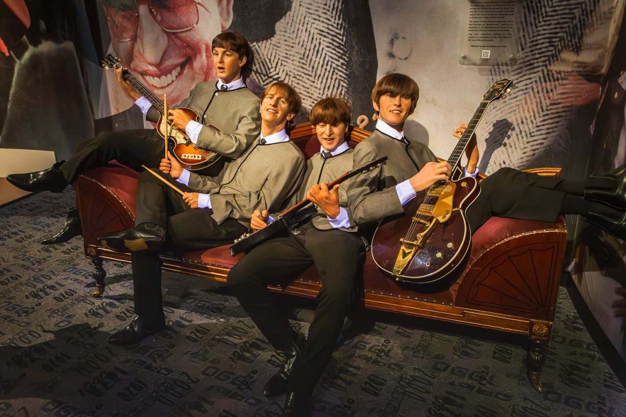 Wax models of the Beatles sitting across a couch in Madame Tussaudes, each holding instruments.