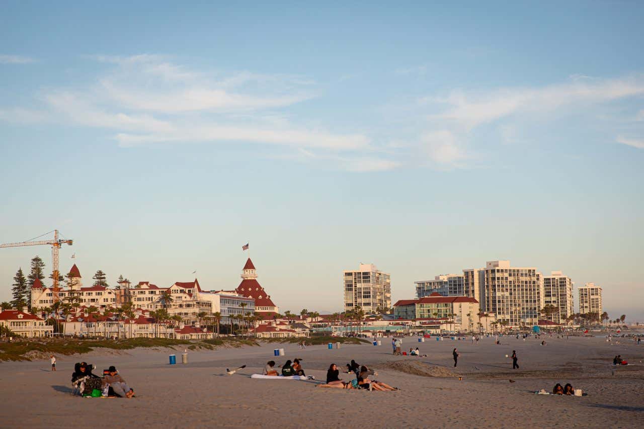 A beach in Coronado with some people sitting on the sand and various buildings in the background.