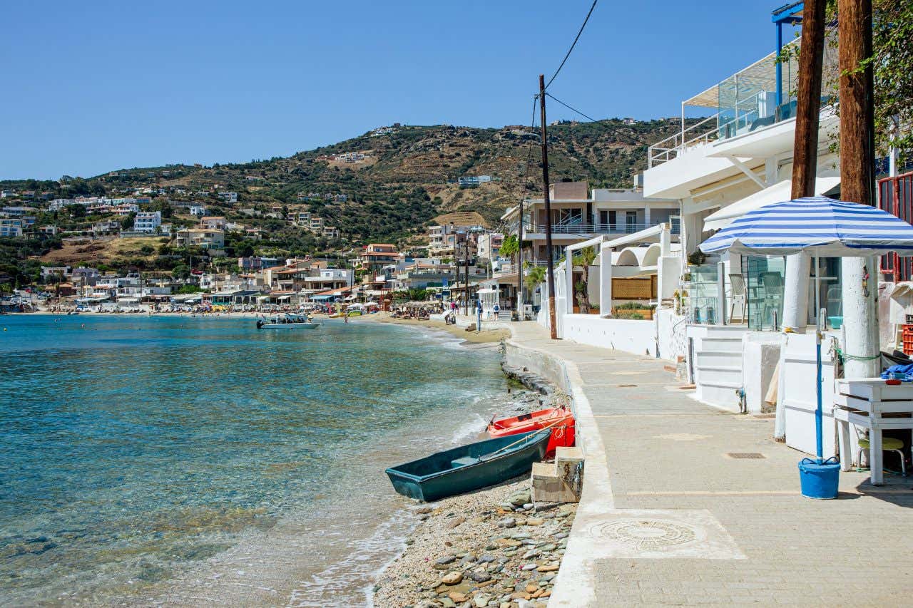 Lungo mare di un piccolo villaggio cretese, con un marciapiede che costeggia la spiaggia fatta di sassi. Sul lato destro dell'immagine, proprio lungo il marciapiede, ci sono dei ristorantini