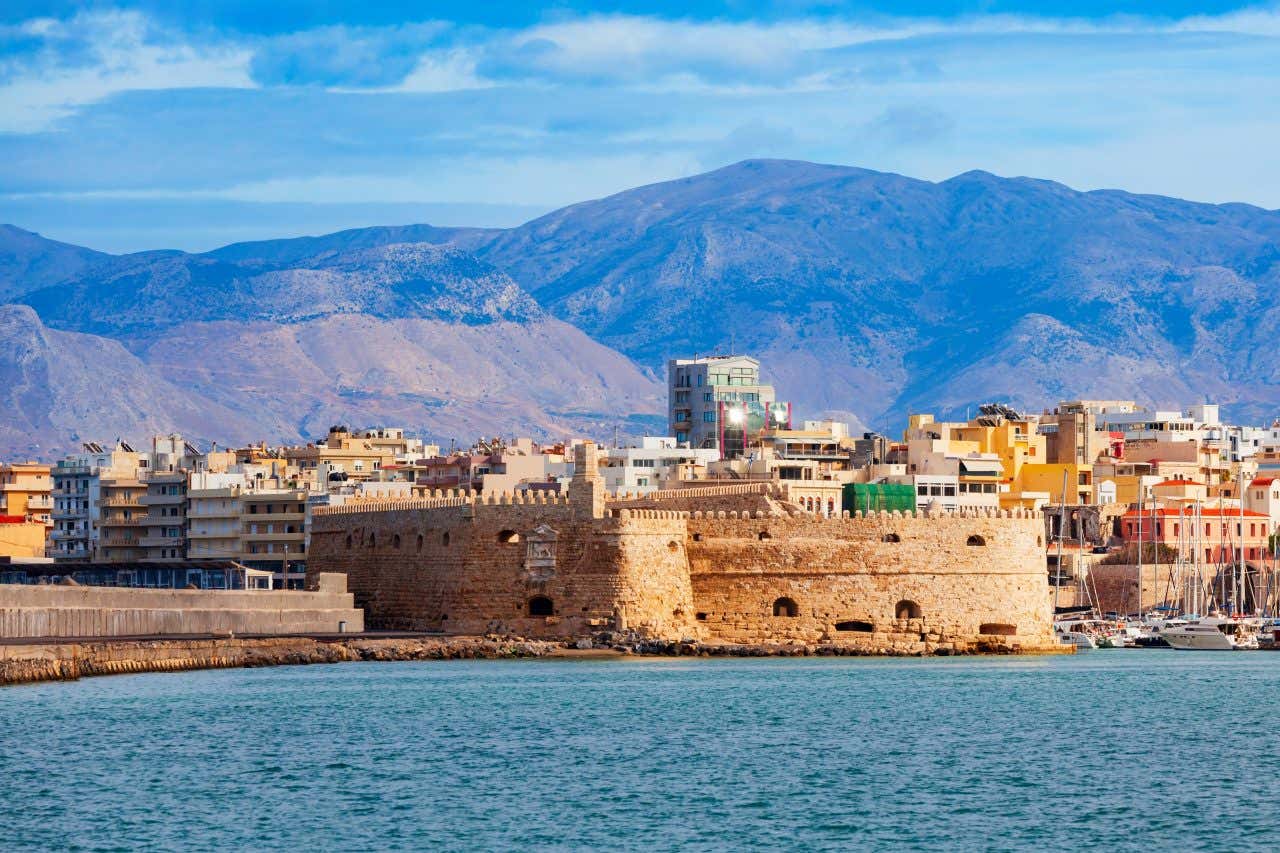 La città di Candia vista dal mare, con una piccola fortezza di pietra bianca in primo piano, palazzi più moderni in secondo piano e alte montagne brulle sullo sfondo
