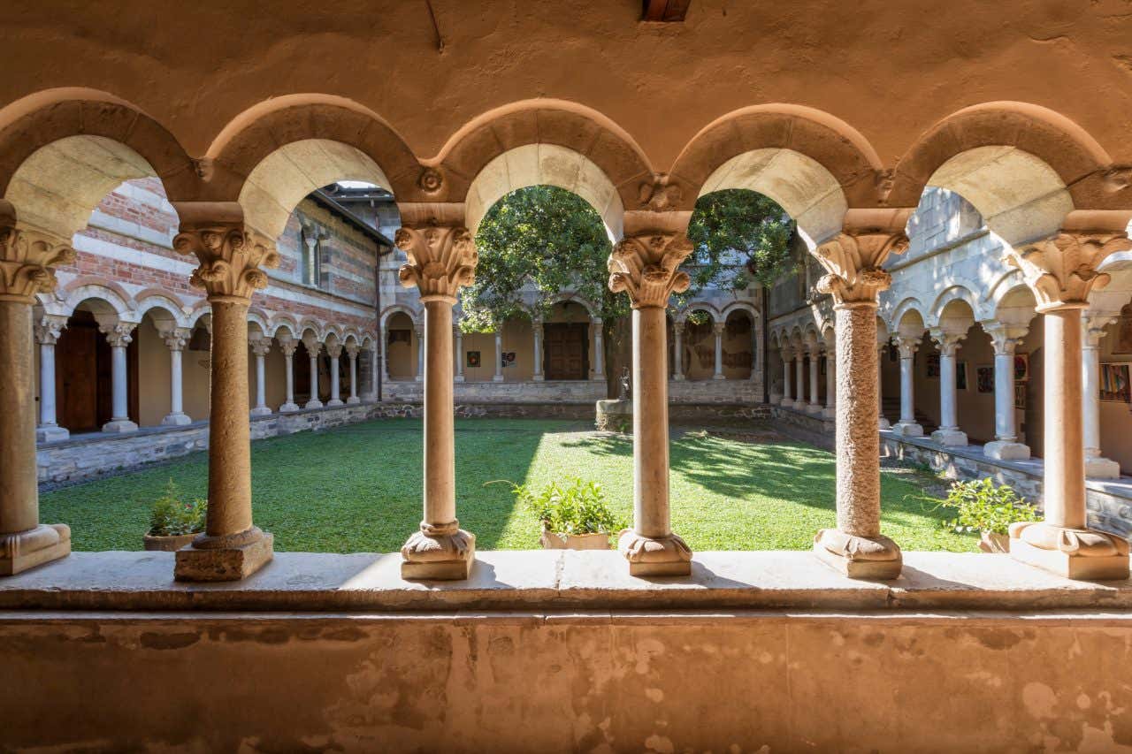 Vista dall'interno del chiostro dell'Abbazia di Piona, altezza uomo, scattata da sotto un'arcata laterale verso il giardino interno, con prato verde ben curato, un antico pozzo di pietra al centro e un albero visibile sul lato in alto a destra