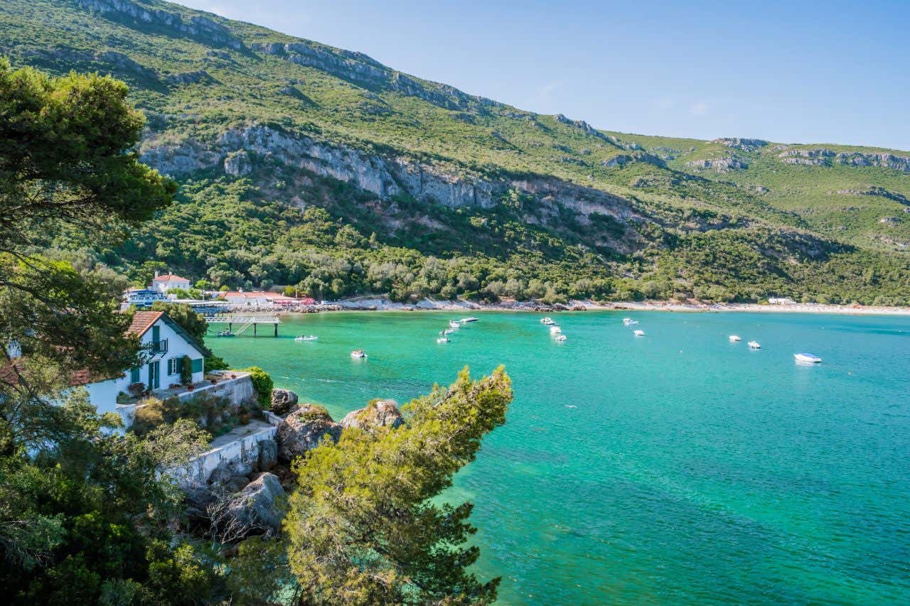 A coastal view of Portinho da Arrábida beach within Arrábida Natural Park, Portugal. It shows a beach with clear turquoise water and green, tree-covered hills. Several small boats are in the water, and a white building is near the shore.