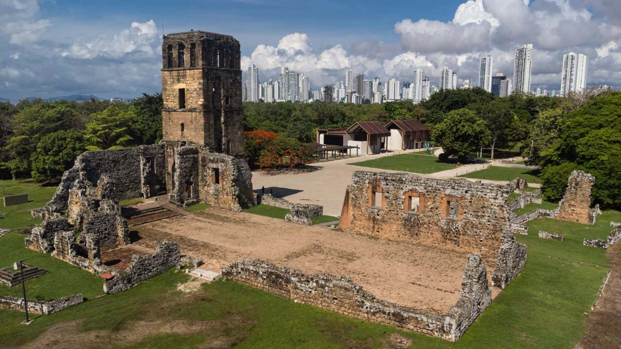 Foto tirada do alto das ruínas do Panamá Viejo, com a alta torre de pedra da igreja e o restante das paredes contra o horizonte da cidade do Panamá e muitas árvores tropicais
