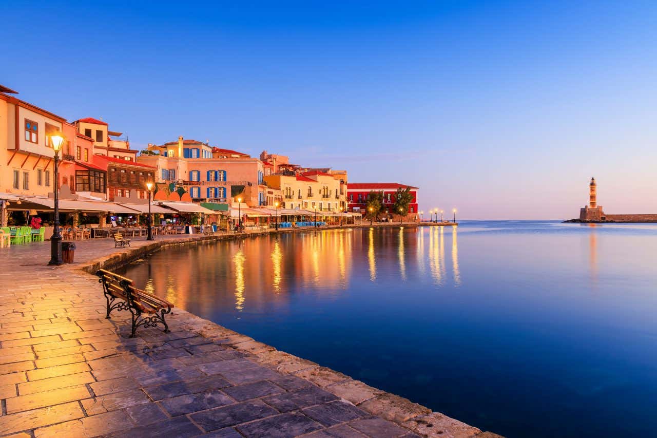 Foto scattata di sera a Chania, Creta. Si vede il lungomare lastricato, illuminato da lampioni in ghisa e decorato con piccole panchine, sullo sfondo un piccolo faro