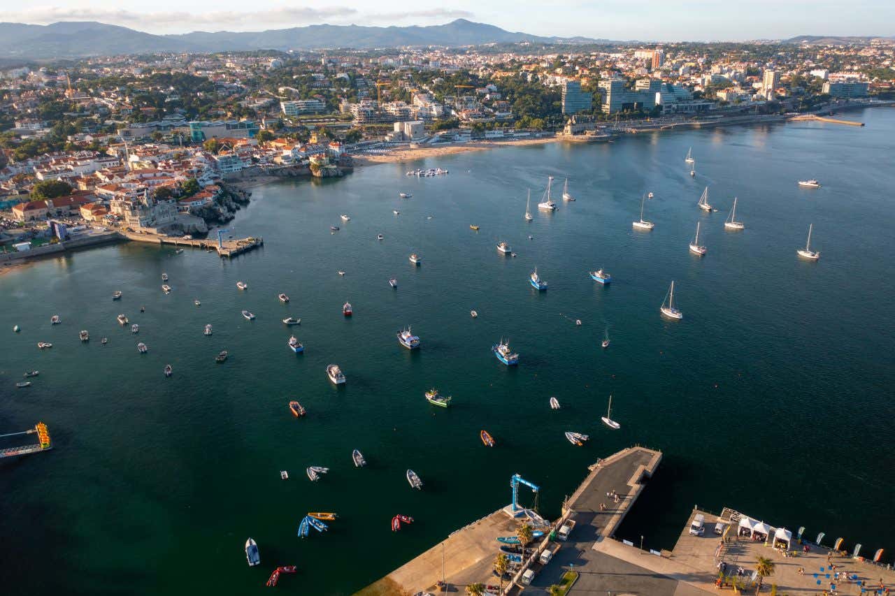 An aerial view of Cascais Marina in Portugal, showing numerous boats in a bay next to a town with buildings along the shoreline. Mountains are visible in the background under a light blue sky.