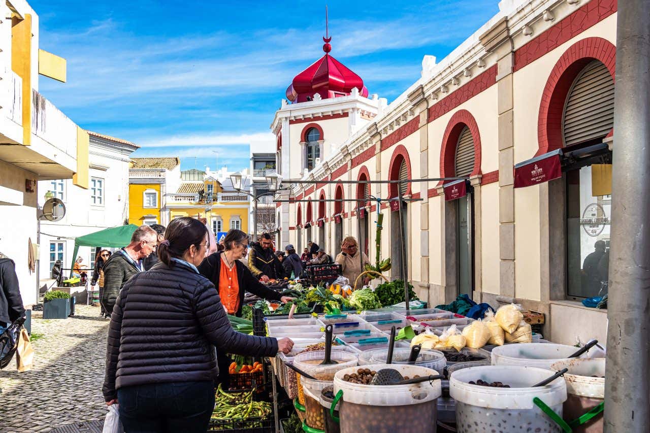 People Browse outdoor market stalls displaying various goods at the Loulé Municipal Market in Portugal. A large building with a distinctive red roof is visible in the background.