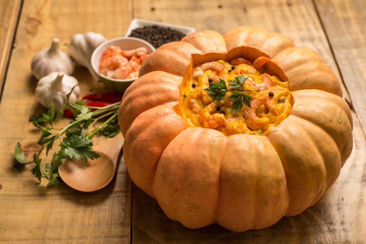 Uma abóbora tipo moranga recheada com creme de camarões em uma mesa de madeira com uma colher de pau, salsinha, pimentas, alho, camarões e pimenta ao lado