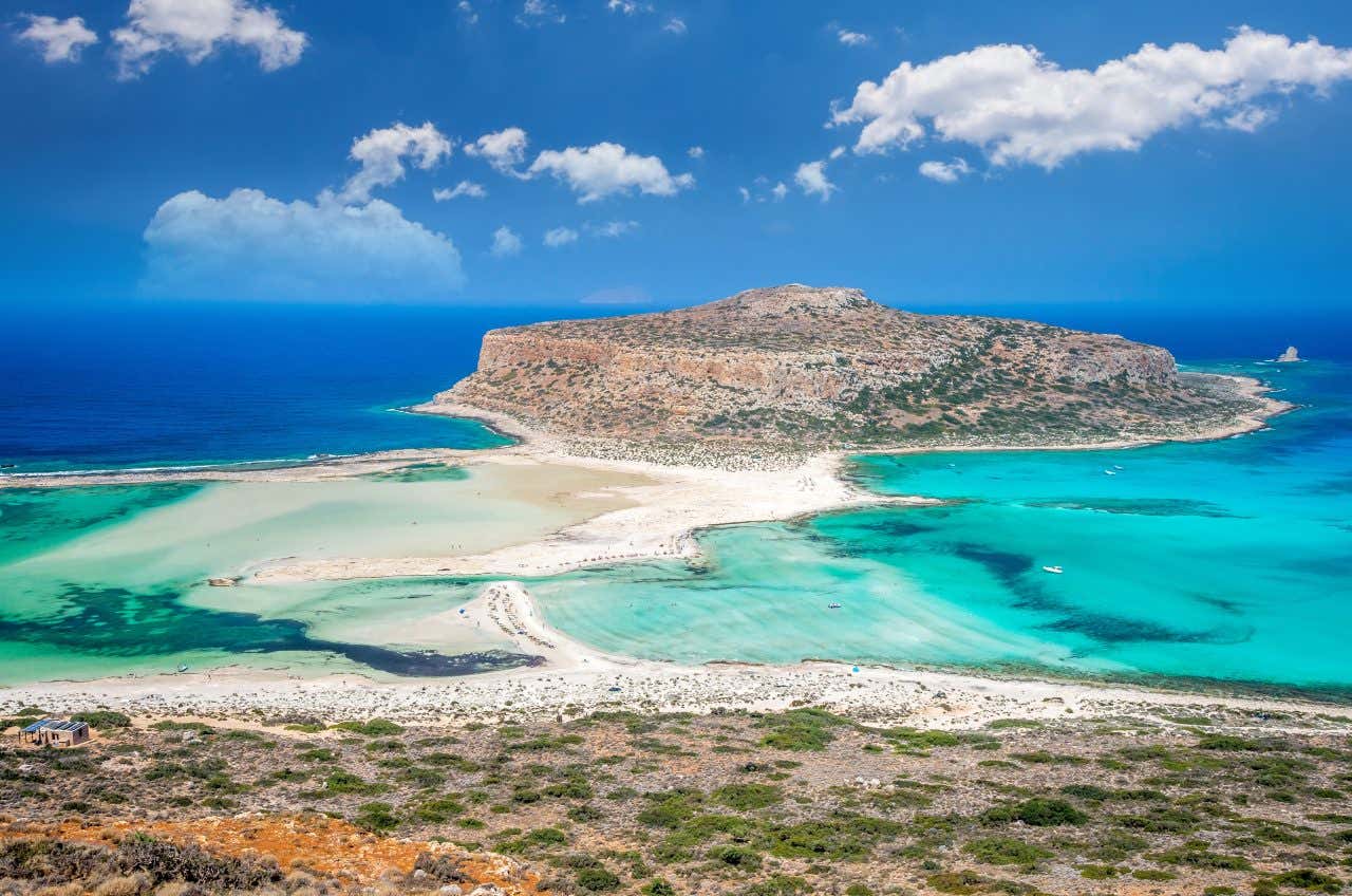 Vista panoramica della laguna di Balos a Creta, Grecia, con acque turchesi, sabbia bianca e un promontorio roccioso circondato dal mare sotto un cielo azzurro con nuvole