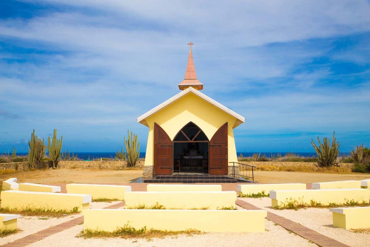 Uma capela amarela com bancos de pedra em frente e o mar do Caribe ao fundo