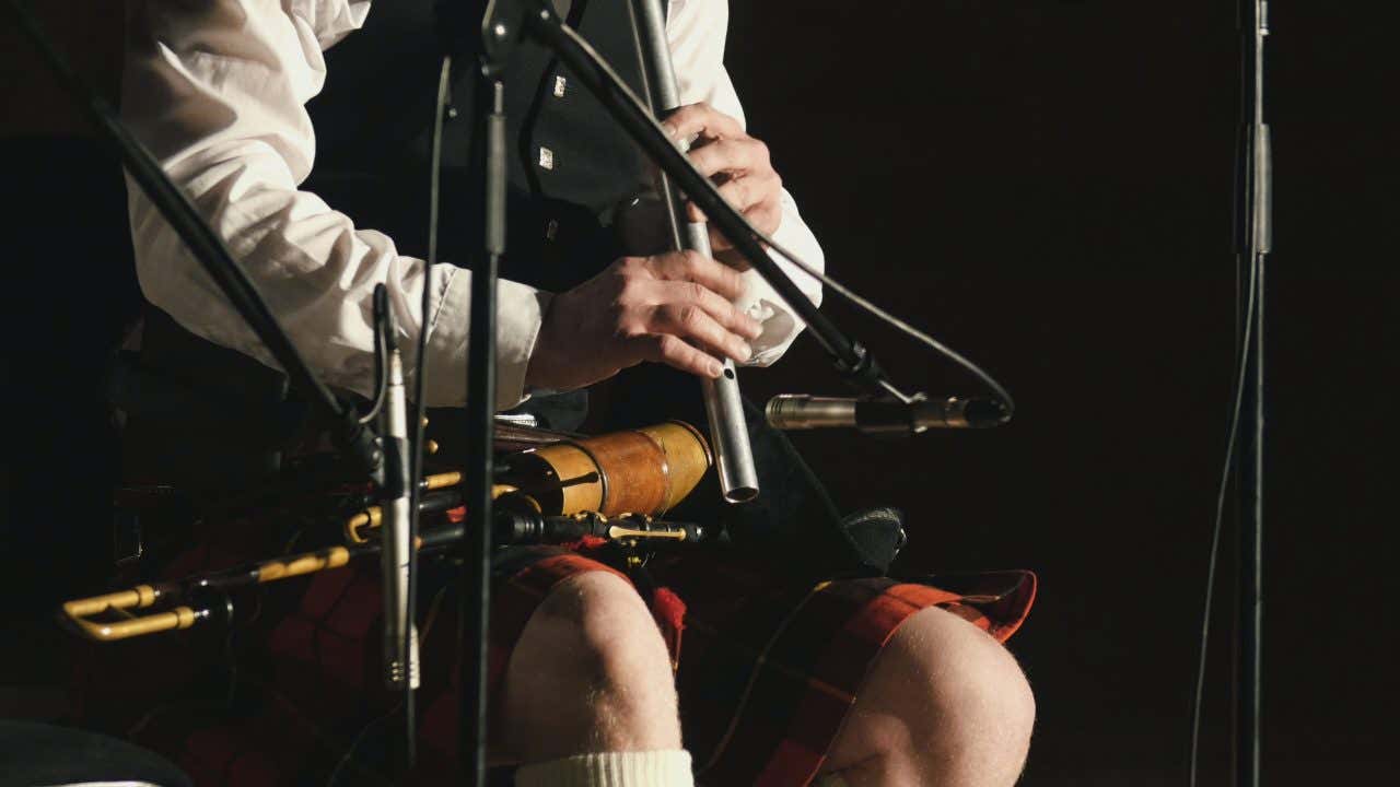 Un musicista scozzese di cui non si vede il volto, è vestito con il classico kilt ed è seduto davanti a un microfono mentre suona un flauto . Sul grembo tiene poggiati quelli che sembrano altri strumenti a fiato