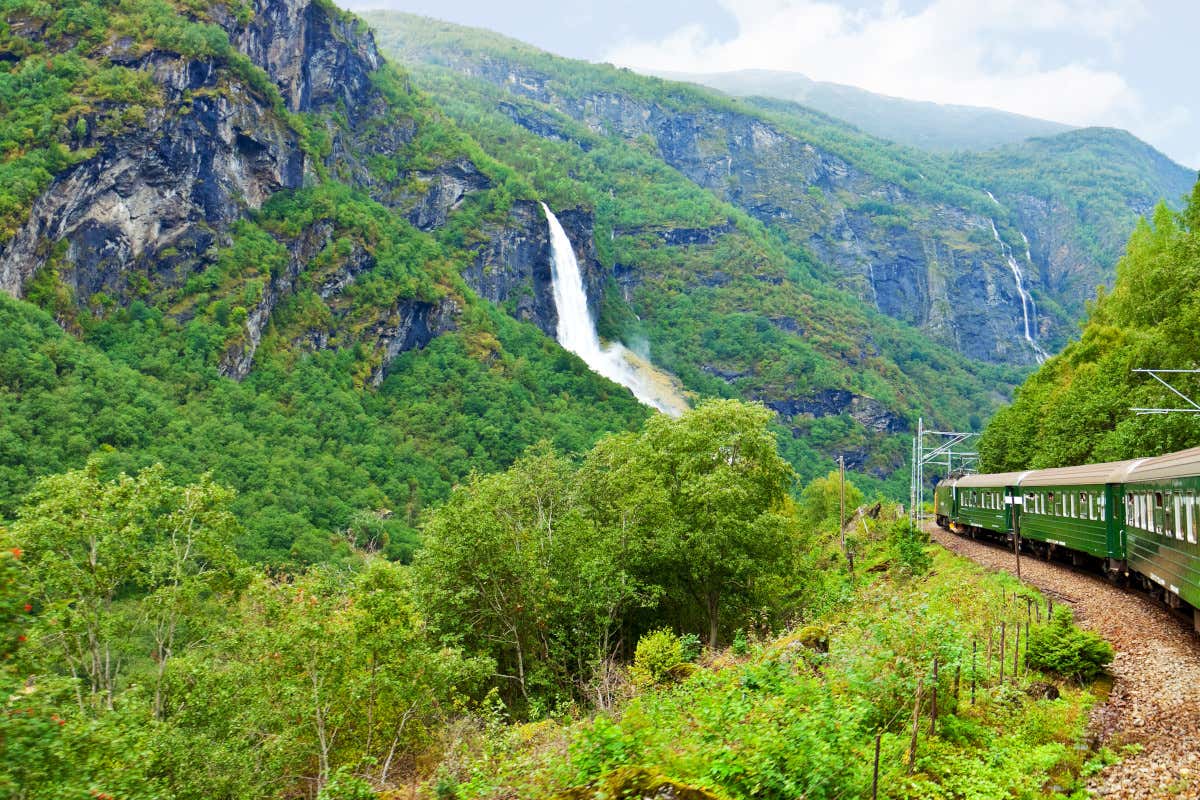 Un tren de color verde atravesando un conjunto de montañas con frondosos árboles y varias cascadas
